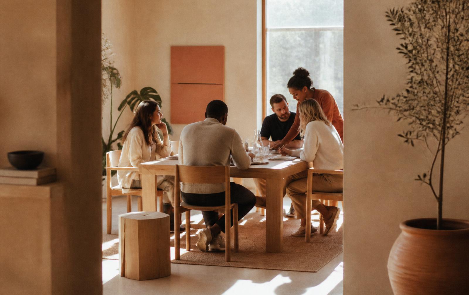 Colleagues discussing project work around a light wooden table in a warm, sunlit room